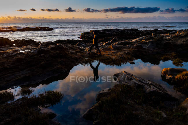 Adolescente caminando en la orilla rocosa casting reflexión bajo el cielo puesta del sol - foto de stock