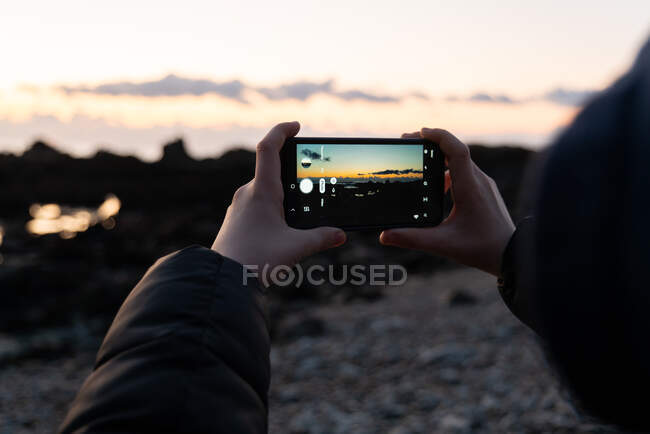 Détail des mains tenant le téléphone intelligent prenant la photo du coucher du soleil sur la plage — Photo de stock