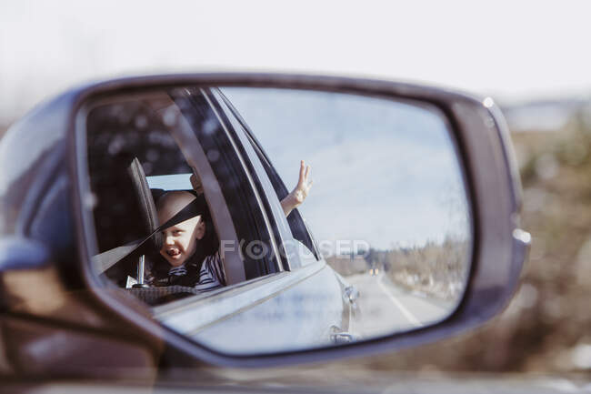 Carro espelho tiro de menino no banco de trás com mão para fora janela no dia ensolarado — Fotografia de Stock