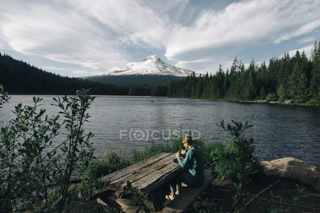 Una joven almorza en la mesa de picnic junto a un lago cerca del monte. Capucha. - foto de stock