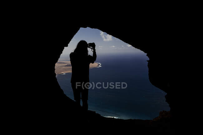 Silueta de un hombre haciendo una foto de una cueva en los acantilados de Famara. - foto de stock