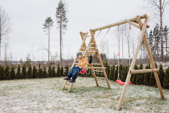 Menino sentou-se em um balanço balançando fora em seu jardim na neve — Fotografia de Stock