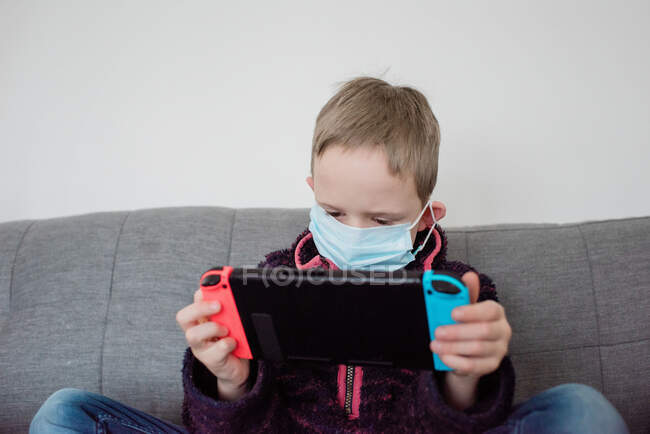 Garçon jouer avec une console de jeu avec un masque facial sur comme protection — Photo de stock
