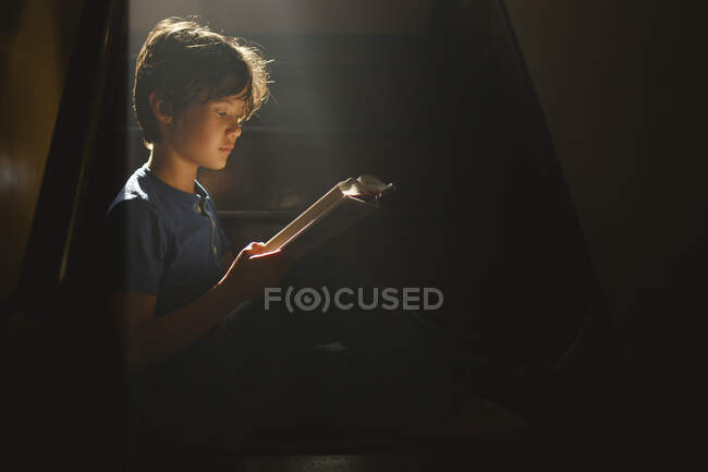 Мальчик сидит на затемненной лестнице в ярком луче света, читая — стоковое фото