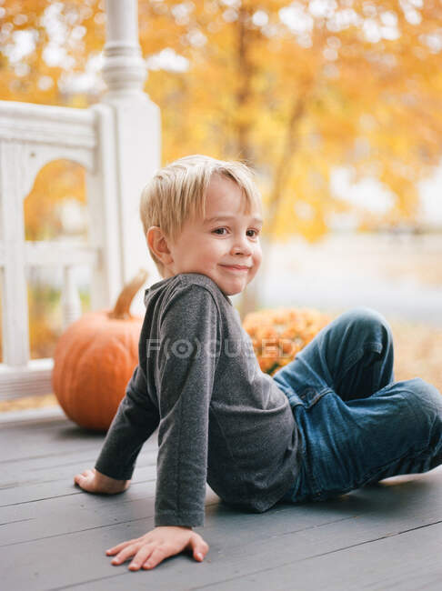 A little boy sitting on a porch by orange maple trees and pumpkins — Stock Photo