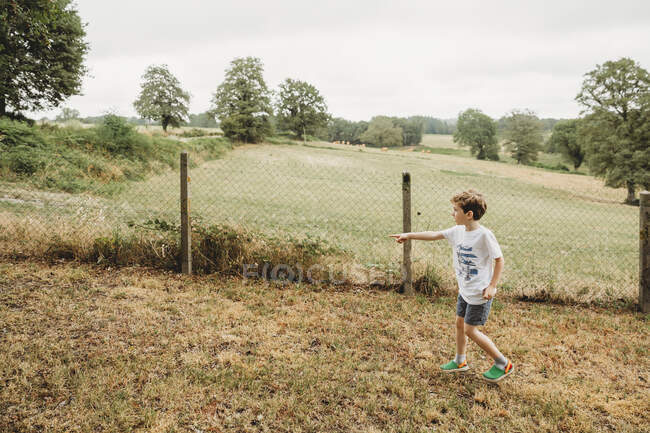 Garçon pointant hors caméra dans le jardin avec des champs en arrière-plan — Photo de stock