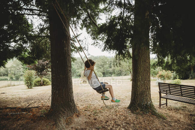 Vista lateral do menino em balanço sob árvores — Fotografia de Stock