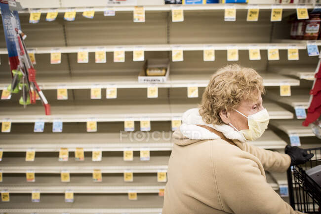 Una mujer con una máscara facial camina junto a estantes vacíos en un supermercado. - foto de stock