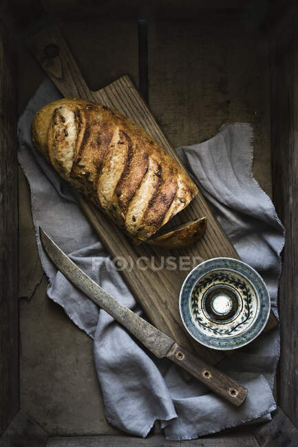 Artisan loaf of bread on board with knife, blue patterned bowl, linen — Stock Photo