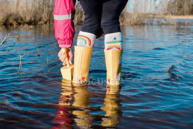 Child's feet in rain boots in the water with a spade playing — Stock Photo