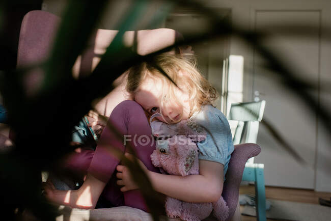 Молода дівчина скручена на стілець, виглядає втомленою, тримаючи свою іграшку вдома — стокове фото