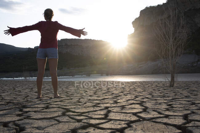 Mujer retroiluminada en el borde de un lago con los brazos extendidos. - foto de stock