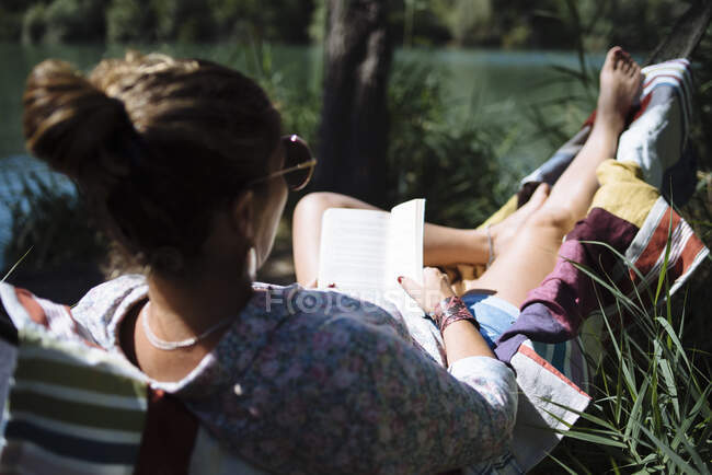 Mulher com óculos de sol lendo um livro deitado em uma rede. — Fotografia de Stock