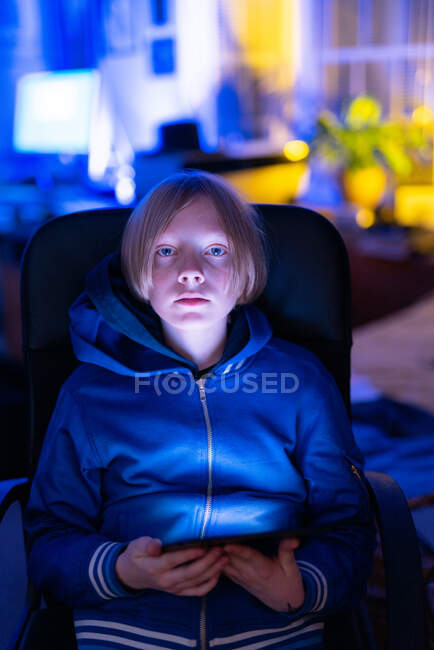 Jeune garçon assis à l'intérieur regardant la caméra tout en travaillant sur tablette — Photo de stock