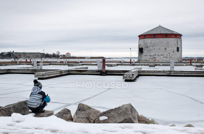 Menino sentado na borda do lago congelado olhando para docas à distância. — Fotografia de Stock