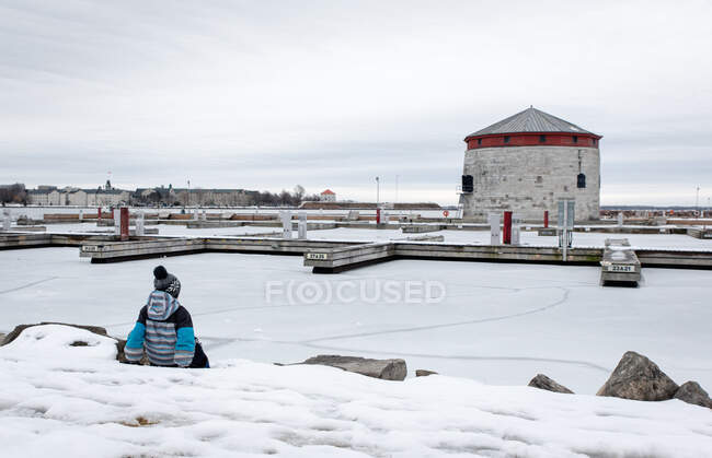 Menino sentado na borda do lago congelado olhando para docas à distância. — Fotografia de Stock