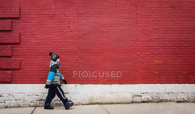 Menino em roupas de inverno andando na calçada na frente da parede vermelha. — Fotografia de Stock