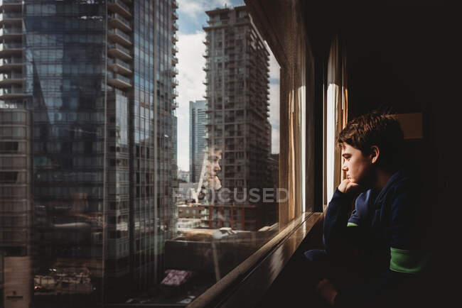 Tween boy looking out a window at tall buildings of the city outside. — Stock Photo