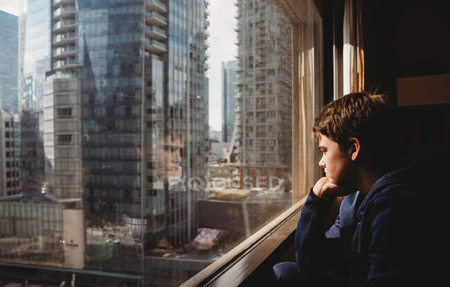 Tween chico mirando por una ventana en edificios altos de la ciudad fuera. - foto de stock