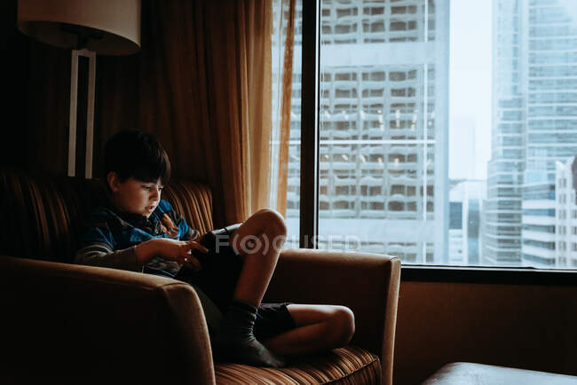 Хлопчик з планшетом у кріслі біля вікна з високими будівлями зовні.. — стокове фото