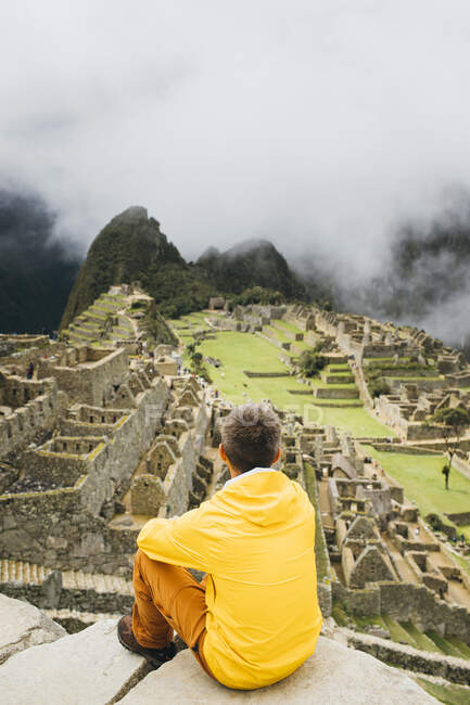 Мужчина в жёлтой куртке сидит возле руин Мачу-Пикчу, Перу — стоковое фото
