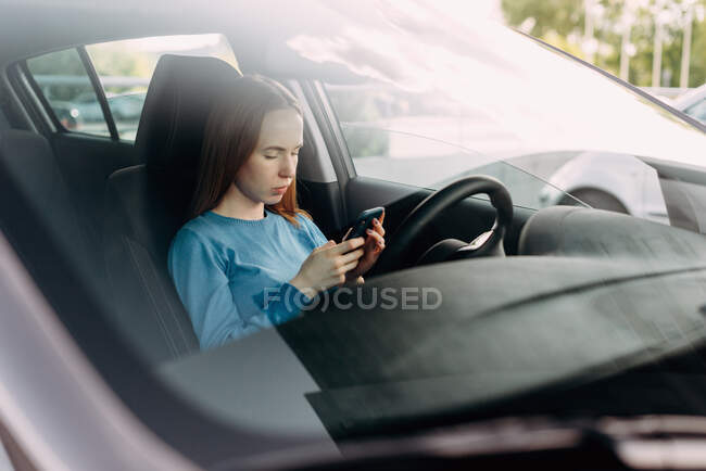 Mujer seria sosteniendo su teléfono inteligente mientras está sentado dentro del coche. - foto de stock