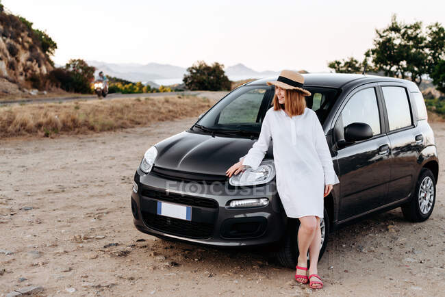 Cute, charming girl in a straw hat poses around a black car on the roadside. — Stock Photo