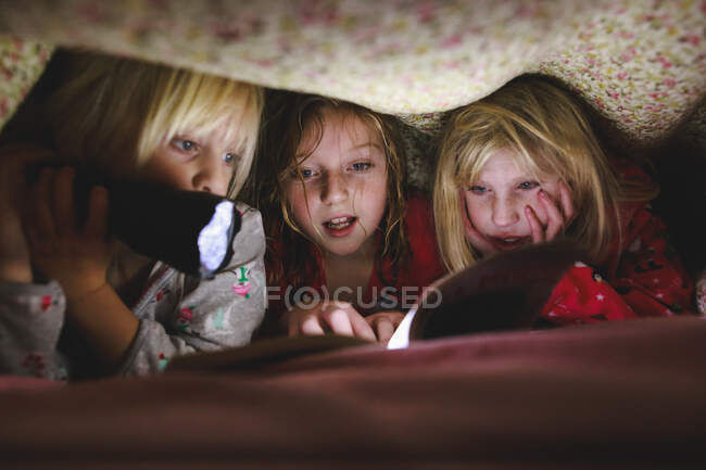Tres niñas leyendo libro bajo manta con linterna - foto de stock