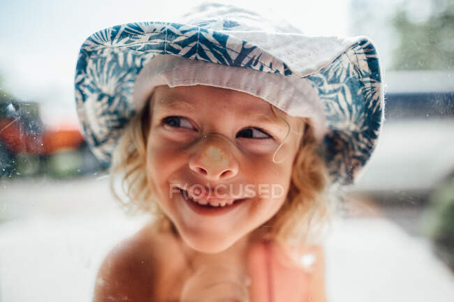 Petit garçon au visage lisse dans une fenêtre portant un chapeau seau — Photo de stock