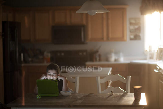 Хлопчик сміється, спостерігаючи за планшетом за кухонним столом, який їсть їжу. — стокове фото