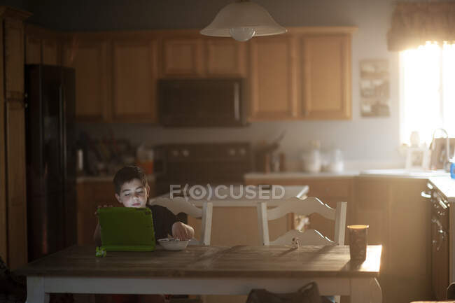 Молодий хлопчик їсть сніданок, спостерігаючи за планшетом на кухні — стокове фото