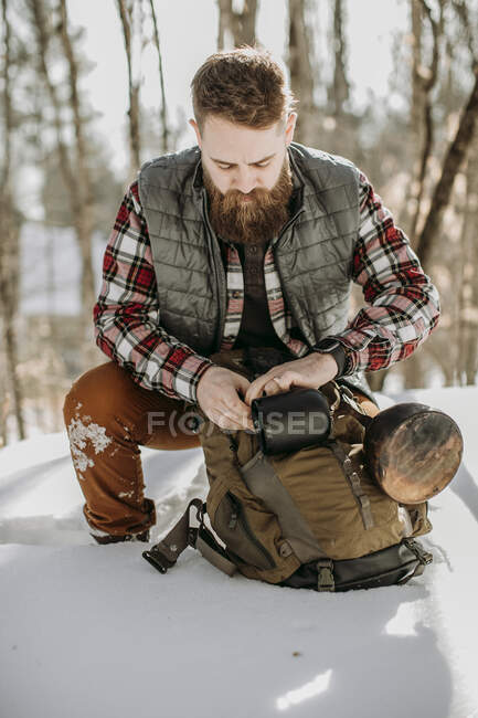 Man with beard wearing flannel kneels in snow and adjusts hiking pack — Stock Photo