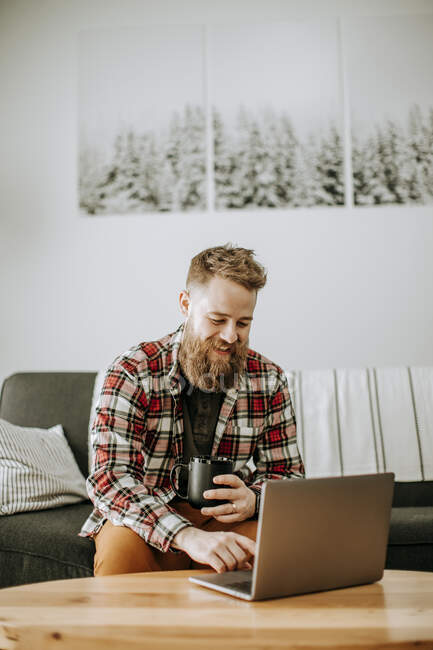 Man with beard holds cup of coffee while working on laptop computer — Stock Photo