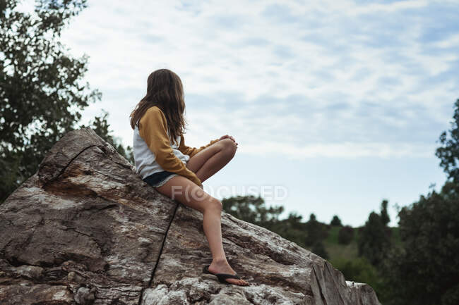 Молода дівчина 10-12 років дивиться на хмари, сидячи на колоді — стокове фото