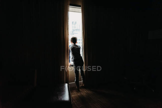 Giovane ragazzo che guarda fuori da una finestra di una stanza buia a edifici alti. — Foto stock