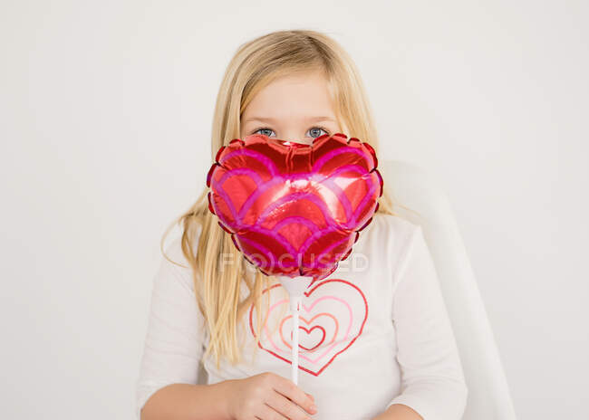 Cute young blond girl holding small red heart balloon under eyes — Stock Photo