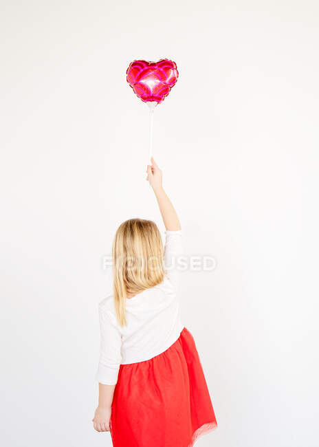 Back view pequena menina loira em tutu vermelho segurando balão coração vermelho — Fotografia de Stock