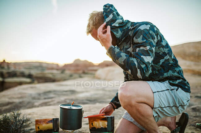 Male camper prepares camp dinner and pulls camo jacket over head — Stock Photo