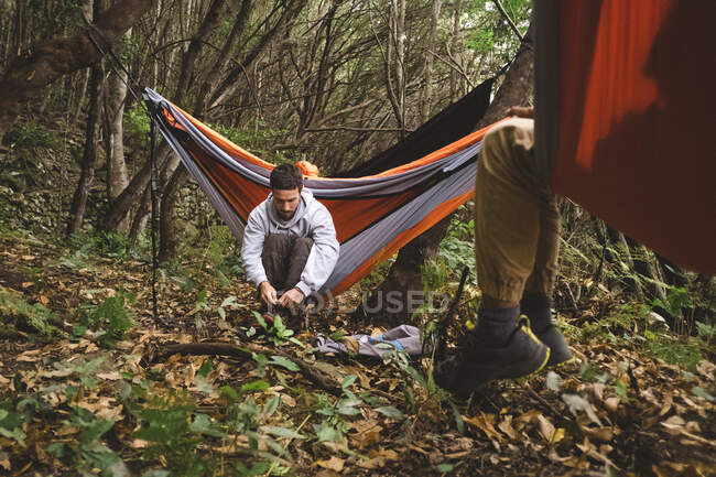 Man sitting in a hammock in the forest gets prepared for hiking — Stock Photo
