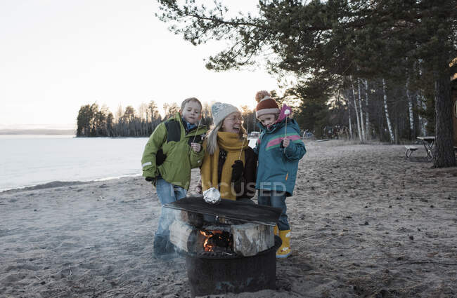 Madre y sus hijos comiendo malvaviscos junto a una fogata en la playa - foto de stock