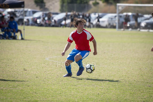 Giocatore di calcio adolescente in uniforme rossa e blu che controlla la palla durante una partita — Foto stock