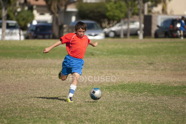 Jeune footballeur en uniforme rouge bleu donnant un coup de pied au ballon de football dans le parc — Photo de stock