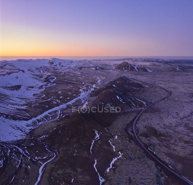 Drohnen-Ansicht einer kurvigen Asphaltstraße, die durch felsiges Gelände mit schneebedeckten Bergen vor dem bunten Sonnenuntergangshimmel in Island verläuft — Stockfoto