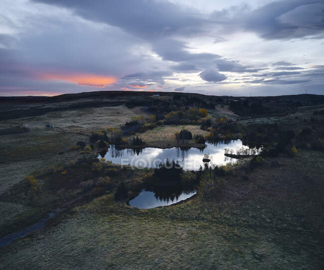 Живописный спокойный пейзаж спокойного маленького озера среди зеленых полей и холмов под облачным закатом неба в исландской сельской местности — стоковое фото