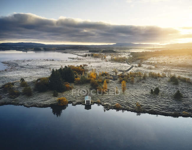 Autunno paesaggio islandese di lago calmo e riva coperta di neve e alberi colorati con casa solitaria situata vicino all'acqua al tramonto con nuvole — Foto stock