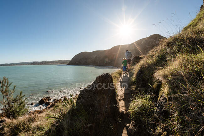 Padre e hijo caminando por un sendero de montaña sobre el océano en Nueva Zelanda - foto de stock