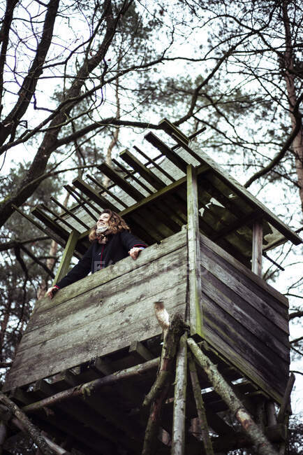 Happy young woman smiles and looks out of high tree house look out — Stock Photo