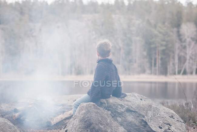 Jeune garçon assis sur un rocher près d'un feu de camp et d'un lac en Suède — Photo de stock