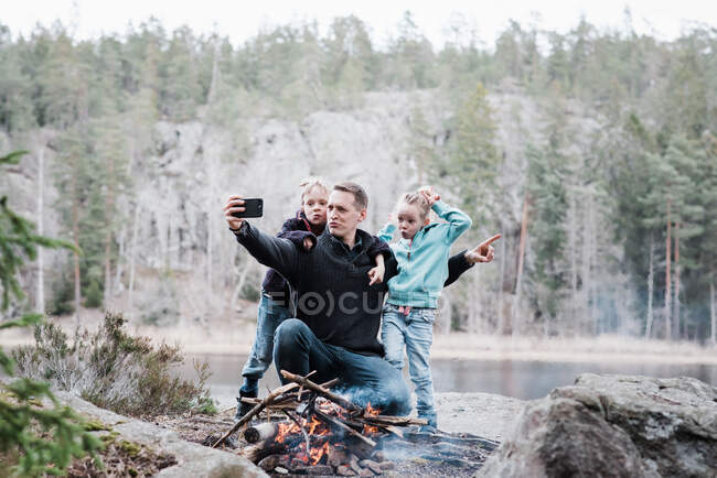 Padre tomando selfies con sus hijos mientras está sentado al lado de una fogata - foto de stock