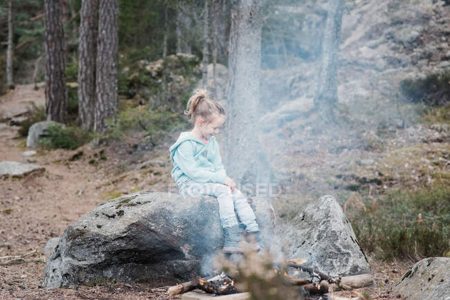 Niña sentada junto a una fogata calentándose en el bosque en Suecia - foto de stock